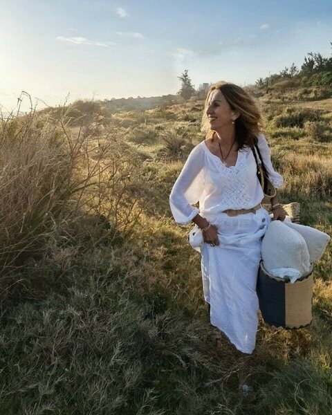 A woman in white walks through tall grass at sunset, carrying a basket of cushions