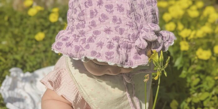 A baby in a flowery hat picks a yellow flower in the grass