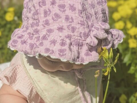 A baby in a flowered hat picks a yellow flower in the grass
